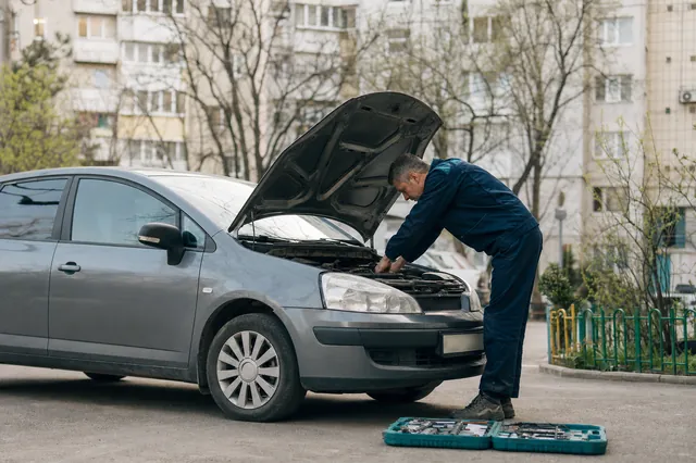 Un mecanic lucrează sub capota unei mașini, în curtea unui bloc